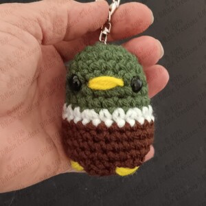 Op de afbeelding: Een gehaakte eend sleutelhanger met een groene kop, bruin lichaam, witte kraag en gele snavel. De eend heeft zwarte ogen en houdt een zilveren sleutelhanger vast.