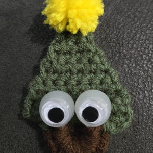 May include: A handmade, whimsical crochet creature with large googly eyes. The body is green, topped with a yellow pom-pom. The base is brown, giving the appearance of a tree or plant-like character. The design is playful and unique.