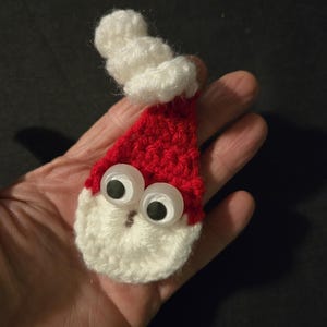 May include: A handmade crochet Santa Claus ornament. The ornament features a red hat with a white pom-pom and beard, and large googly eyes. The Santa Claus is held in a hand against a dark background.