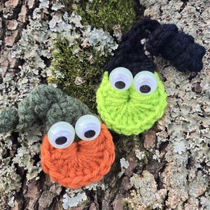 Peut inclure: Deux créatures faites à la main au crochet avec des yeux globuleux reposent sur un tronc d'arbre recouvert de mousse. L'une est orange avec un chapeau vert, l'autre est vert citron avec un chapeau noir. Les figurines fantaisistes sont probablement décoratives.