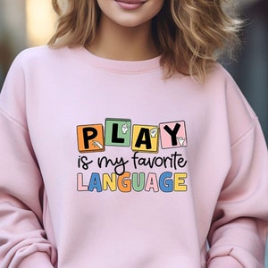 May include: Pink sweatshirt with the text "PLAY is my favorite LANGUAGE" in colorful blocks with hand signs in each block.