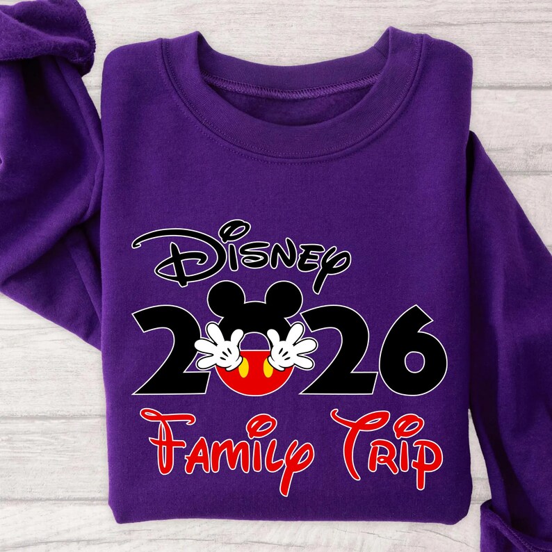K&ouml;nnte beinhalten: Ein lila Sweatshirt mit dem Wort "Disney" &uuml;ber den Zahlen "2026", wobei das Gesicht von Mickey Mouse in die Null integriert ist. Die Worte "Family Trip" stehen darunter in roter Schrift.