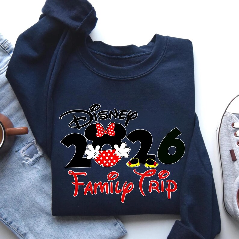 K&ouml;nnte beinhalten: Marineblaues Sweatshirt mit Minnie Mouse-Design und dem Text "Disney 2026 Family Trip" in Rot und Schwarz. Das Design beinhaltet Minnie's Schleife und gepunkteten Rock. Ein l&auml;ssiges Oberteil f&uuml;r den Familienurlaub.