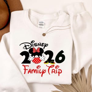 K&ouml;nnte beinhalten: Wei&szlig;es Sweatshirt mit einem Disney 2026 Family Trip-Design. Das Design zeigt Minnie Mouse mit einem roten gepunkteten Rock und Schleife, sowie den Text "Disney" in schwarzer Schrift &uuml;ber den Zahlen "2026" und den Worten "Family Trip" in roter Schrift.