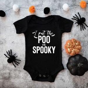 K&ouml;nnte beinhalten: Ein schwarzer, kurz&auml;rmeliger Babybody mit dem wei&szlig;en Schriftzug I put the POO in SPOOKY und kleinen Fledermausgrafiken. Das Kleidungsst&uuml;ck hat einen Rundhalsausschnitt und drei Druckkn&ouml;pfe aus Metall im Schrittbereich zum Verschlie&szlig;en.