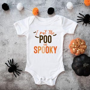 K&ouml;nnte beinhalten: Ein wei&szlig;er kurz&auml;rmeliger Babybody mit dem Schriftzug "I put the POO in SPOOKY" in braunen und orangefarbenen Buchstaben, verziert mit kleinen schwarzen Fledermausgrafiken. Das Kleidungsst&uuml;ck hat einen Rundhalsausschnitt und drei Druckkn&ouml;pfe.