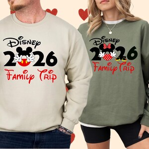K&ouml;nnte beinhalten: Zwei lang&auml;rmlige Sweatshirts, eines beige und eines olivgr&uuml;n, mit einem Disney-Design. Das Design enth&auml;lt den Text "Disney 2026 Family Trip" mit Mickey Mouse-Bildern. Das beige Sweatshirt ist links, das gr&uuml;ne rechts.