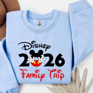 K&ouml;nnte beinhalten: Hellblaues Sweatshirt mit dem Wort "Disney" in schwarzer Schrift &uuml;ber der Zahl "2026" mit Mickey Mouse Ohren und H&auml;nden. Die Worte "Family Trip" stehen in roter Schrift darunter.