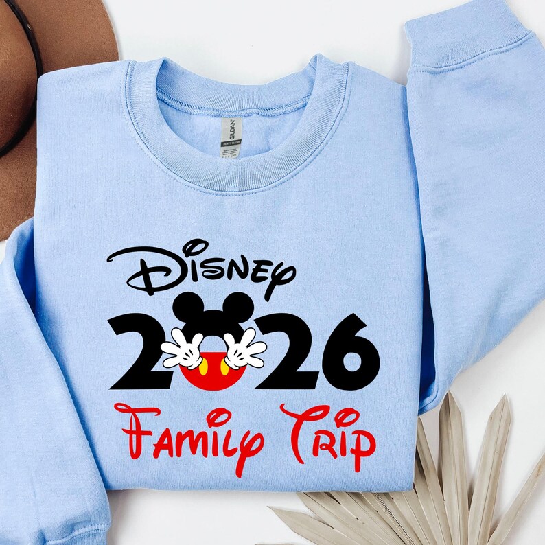 K&ouml;nnte beinhalten: Hellblaues Sweatshirt mit dem Wort "Disney" in schwarzer Schrift &uuml;ber der Zahl "2026" mit Mickey Mouse Ohren und H&auml;nden. Die Worte "Family Trip" stehen in roter Schrift darunter.