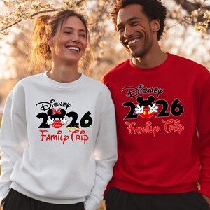 K&ouml;nnte beinhalten: Zwei Personen tragen passende Disney-Sweatshirts. Eines ist wei&szlig;, das andere rot. Beide haben das Disney-Logo, die Zahl 2026 und die Aufschrift "Family Trip" aufgedruckt. Das Design beinhaltet Micky und Minnie Maus.