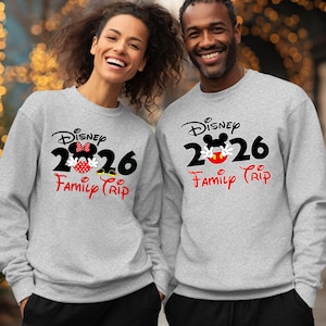 K&ouml;nnte beinhalten: Zwei Personen tragen passende graue Sweatshirts mit einem Disney-Design. Das Design enth&auml;lt das Wort "Disney", das Jahr "2026" und die Worte "Family Trip" in Rot. Ein Sweatshirt zeigt Minnie Mouse, das andere Mickey Mouse.