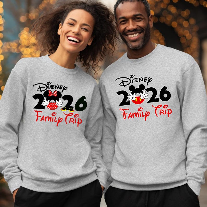 K&ouml;nnte beinhalten: Zwei Personen tragen passende graue Sweatshirts mit einem Disney-Design. Das Design enth&auml;lt das Wort "Disney", das Jahr "2026" und die Worte "Family Trip" in Rot. Ein Sweatshirt zeigt Minnie Mouse, das andere Mickey Mouse.