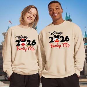 K&ouml;nnte beinhalten: Zwei beige Sweatshirts mit dem Disney-Logo, der Zahl 2026 und den Worten "Family Trip" in Rot. Ein Sweatshirt hat ein Minnie Mouse-Design, das andere ein Mickey Mouse-Design. Die Sweatshirts werden von zwei Personen vor einem Schloss getragen.