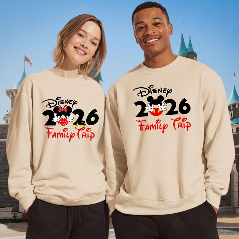 K&ouml;nnte beinhalten: Zwei beige Sweatshirts mit dem Disney-Logo, der Zahl 2026 und den Worten "Family Trip" in Rot. Ein Sweatshirt hat ein Minnie Mouse-Design, das andere ein Mickey Mouse-Design. Die Sweatshirts werden von zwei Personen vor einem Schloss getragen.