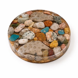 Könnte beinhalten: Ein runder Untersetzer aus Kunstharz mit einer Ansammlung farbiger Steine und Kieselsteine. Die Steine variieren in Größe, Form und Farbe, darunter Beige-, Orange-, Blau- und Grautöne. Der Untersetzer hat einen goldfarbenen Rand.