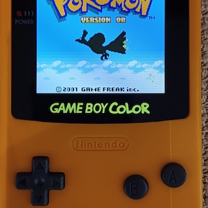 May include: An orange Game Boy Color handheld console displaying the Pokémon Version OR game. The screen shows a blue sky with a silhouette of a bird. The console has black buttons and the text "GAME BOY COLOR".
