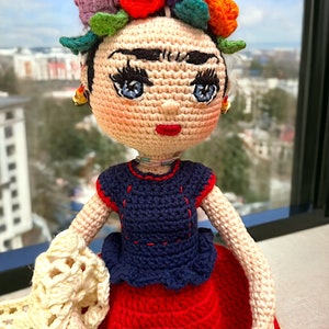May include: A crocheted doll wearing a colourful flower crown, blue eyes, and a red and blue dress. The doll is holding a white crocheted shawl.