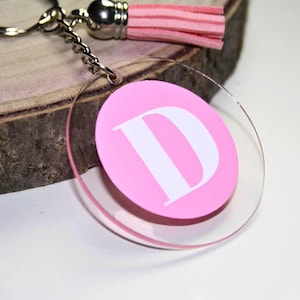Può includere: Portachiavi in acrilico rosa con una lettera D bianca su sfondo trasparente. Il portachiavi ha un anello portachiavi argentato e un pon pon rosa.