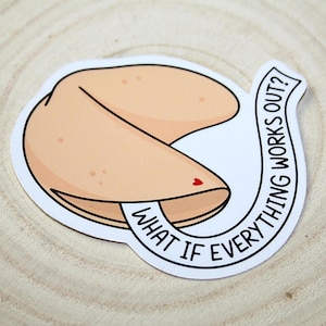 May include: A sticker featuring a fortune cookie design with the text "WHAT IF EVERYTHING WORKS OUT?" in a circular banner. The fortune cookie is light brown with a small red heart detail.
