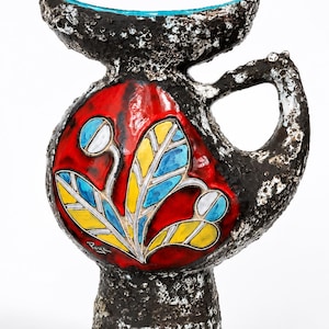 Peut inclure: Un pichet en céramique avec un extérieur texturé brun foncé et noir à la surface rugueuse et cratérisée. L'avant présente un médaillon rouge lisse et brillant avec un motif floral peint en jaune, bleu et blanc. Le bord intérieur est bleu vif.