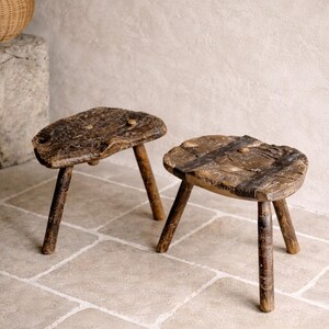May include: Two rustic wooden stools, each with three legs. The seats are irregularly shaped and made from reclaimed wood. The stools are dark brown and have a natural, aged appearance. The stools are approximately 30 cm in height.