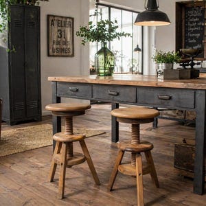 Op de afbeelding: Een rustieke keuken met een grote houten tafel met een zwart metalen frame en drie laden. Twee verstelbare houten krukken met schroefmechanismen staan voor de tafel. Een vintage bord leest "ATELIER 31 RUE DES USINES."