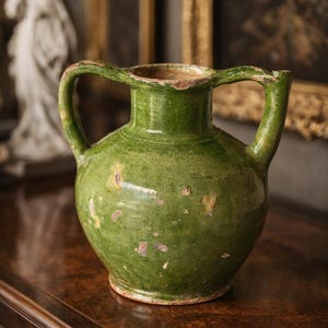 Peut inclure: Une cruche en céramique verte à deux anses. La cruche a un corps arrondi et un bord légèrement évasé. La surface présente un aspect moucheté avec des zones de couleur plus claire. La cruche est posée sur une surface en bois sombre.