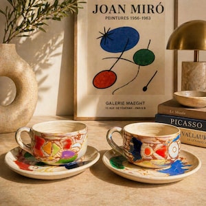 Peut inclure: Deux tasses et soucoupes en céramique avec des motifs abstraits colorés. Les tasses et les soucoupes ont une base de couleur crème avec des accents rouges, bleus, verts et jaunes. L'arrière-plan comprend une impression d'art de Joan Miró et des livres.