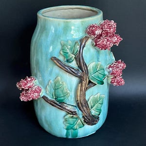 May include: A cylindrical ceramic vase with a glossy light blue glaze. The exterior features a raised, three-dimensional relief of a brown tree branch with green leaves and clusters of textured, deep red flowers extending from the sides.
