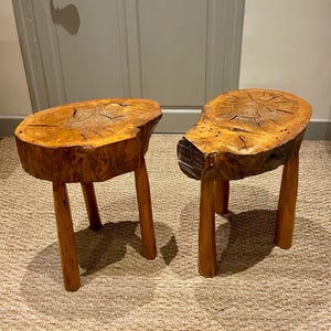 May include: Two rustic wooden stools. Each stool has a natural, irregular-shaped top with visible wood grain and cracks. The tops are supported by three sturdy, rounded legs. The stools are made of light brown wood.