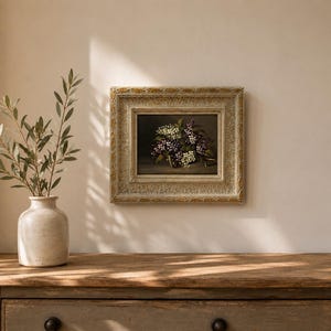 Peut inclure: Une peinture à l'huile encadrée représentant un bouquet de lilas violets et blancs sur fond sombre, placée dans un cadre en bois crème vieilli et orné de détails dorés, exposée sur un mur au-dessus d'une surface en bois.