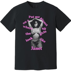 May include: A black short-sleeved t-shirt featuring a central black and white photographic print of a person with hands covering their chest. The text 'Put me on a planet where all the girls look like Janet' is printed in pink around the image.