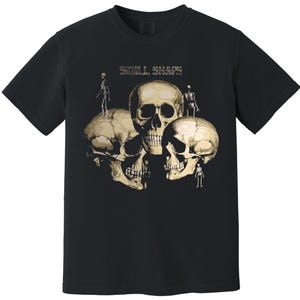 Puede incluir: Camiseta negra con un gráfico de tres calaveras en tonos beige y crema, con pequeñas figuras esqueléticas. El texto "SKULL SHAPES" está impreso encima de las calaveras. La camiseta tiene cuello redondo.