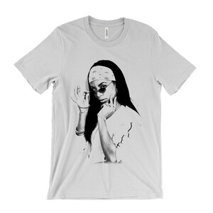 May include: A light gray t-shirt featuring a black and white graphic of a person wearing sunglasses and a bandana. The person is touching their face and sunglasses. The shirt is a crew neck style.