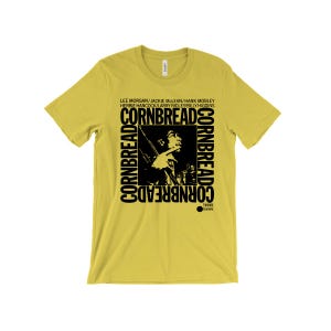 Puede incluir: Una camiseta amarilla con un estampado gráfico en blanco y negro de un músico tocando el saxofón. El estampado gráfico incluye el texto "CORNBREADS" repetido varias veces. El texto "LEE MORGAN/JACKIE MCLEAN/HANK MOBLEY HERBIE HANCOCK/LARRY RIDLEY/BILLY HIGGINS" está impreso sobre el gráfico.