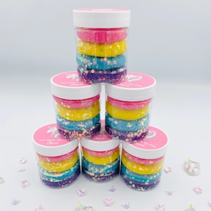 Peut inclure: Une pile de cinq pots transparents remplis de slime coloré. Chaque pot a un couvercle rose et une étiquette qui dit "You Make My Day Magical". La slime est en couches de rose, jaune, bleu et violet.