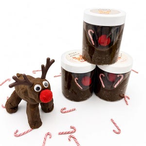 May include: A brown modeling clay reindeer figure with googly eyes, a red pom-pom nose, and brown antlers, positioned next to three clear plastic jars filled with brown dough, each topped with a white lid labeled Build A Reindeer.