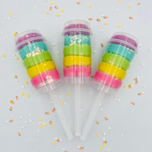 May include: Three rainbow-colored push pop containers filled with layers of pink, yellow, green, and teal dough, topped with purple and glitter. Each container has a clear plastic stick and lid. The background is white with confetti.