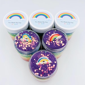 May include: Five clear jars filled with colorful layered slime, each topped with a rainbow charm and purple glitter. The jars have white lids with rainbow and cloud designs and the text "You Made My Day Bright".