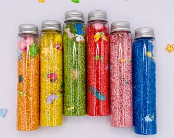 May include: Seven clear glass bottles filled with colorful rice and small plastic toys. The bottles are arranged in a row from left to right, with the colors of the rice progressing from orange to yellow, green, red, pink, and blue.