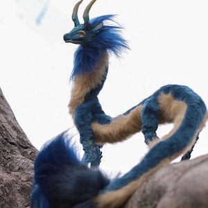 May include: A detailed plush dragon sculpture with a blue and tan color scheme. The dragon has curved horns, a long neck, and a flowing tail. The dragon is posed on a tree branch against a white background.