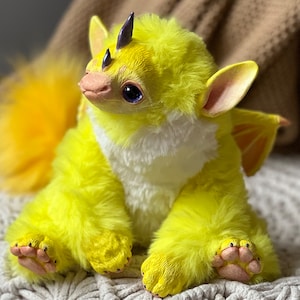 May include: A plush, yellow fantasy creature with a fluffy tail, wings, and small horns. The creature has a white chest, pink paws, and large, dark eyes. It is sitting on a white, textured surface.