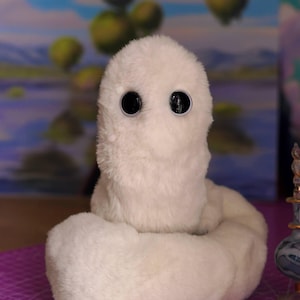 May include: A plush white creature with a soft, fuzzy texture and two large, round, black eyes. The figure has a rounded head and a coiled, thick body. The text "Edition: Artist made" is printed in white in the bottom right corner of the image.