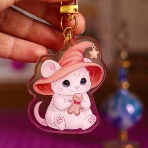 Peut inclure: Un porte-clés en acrylique représentant un chaton blanc portant un grand chapeau de magicien rose avec une petite breloque en forme d'étoile. Le chaton a un croissant de lune sur le front et tient un petit biscuit bonhomme en pain d'épices.
