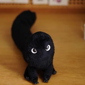 Könnte beinhalten: Ein Plüschtier in Form eines langen, segmentierten Wesens, das mit weichem schwarzem Fell bedeckt ist. Es hat zwei große, runde weiße Augen mit schwarzen Pupillen an der Vorderseite des Kopfes und vier kleine, dunkle Gliedmaßen am Unterkörper.