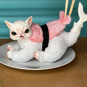 May include: A whimsical plush cat sushi roll on a white plate. The white cat has green eyes and pink paws. It is adorned with a pink salmon-like topping and a black seaweed belt. Chopsticks are positioned above the tail.
