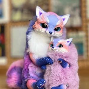 Könnte beinhalten: Zwei Plüschfuchsfiguren, eine große und eine kleine, in einer Umarmung. Die Füchse haben blaues, rosa und lila Fell mit weißen Akzenten. Der größere Fuchs hat einen langen, flauschigen Schwanz. Der kleinere Fuchs ist in den Armen des größeren Fuchses eingebettet.