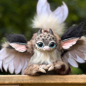 Puede incluir: Una muñeca de criatura fantástica hecha a mano con una cabeza manchada, ojos azules grandes y alas de plumas. La muñeca tiene un cuerpo esponjoso, marrón y beige con acentos negros.