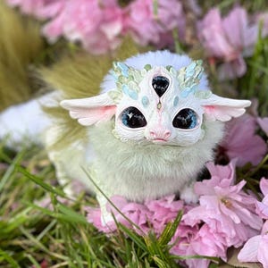 May include: A fantastical, white creature with three eyes and large, blue pupils. It has fluffy, light green fur and delicate, pink-lined ears. Adorned with a crown of blue and green leaves, it rests amidst pink blossoms and green grass.