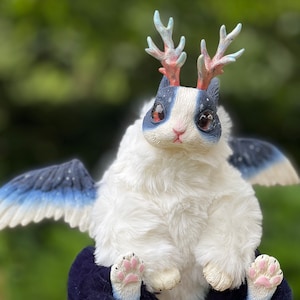 May include: A fantastical plush toy with white fur, blue and white wings, and blue and white facial markings. It features small antlers and pink paw pads. The creature is seated on a dark blue base.